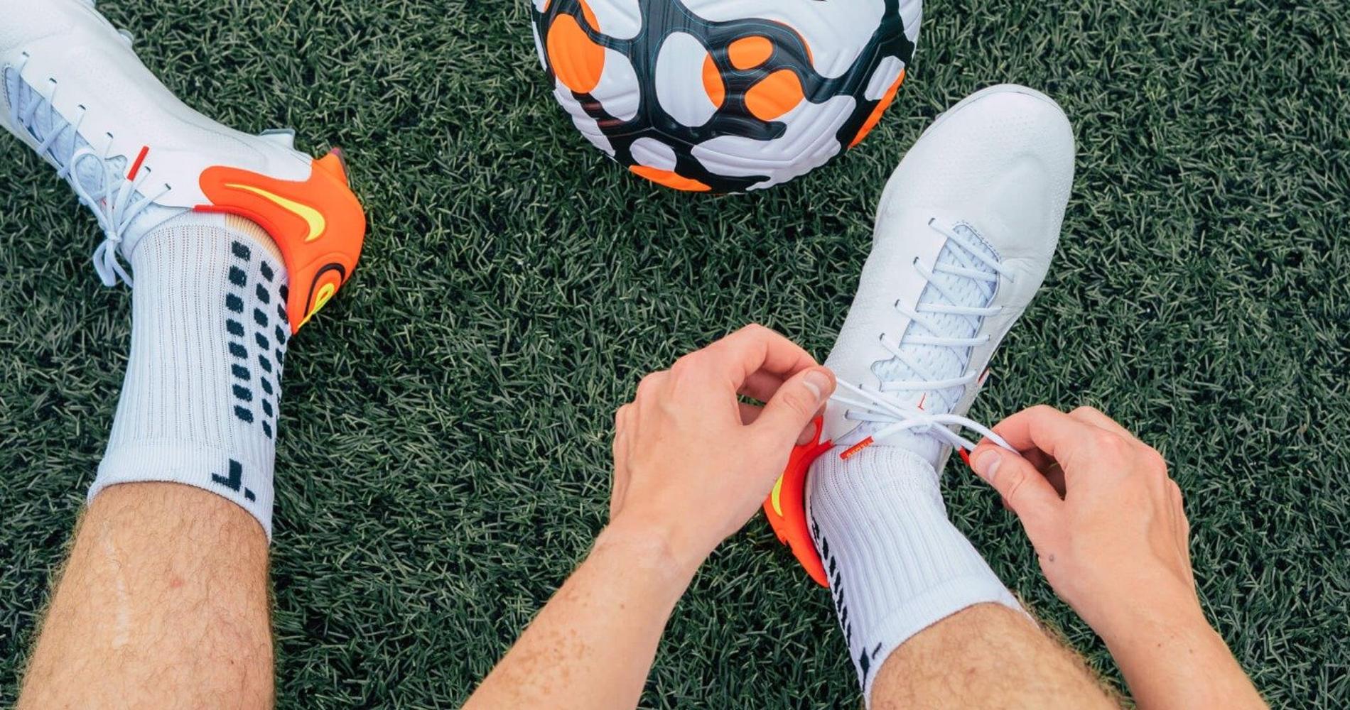 Joueur de football nouant ses crampons avec des chaussettes antidérapantes blanches NoGlide Performance sur gazon synthétique.